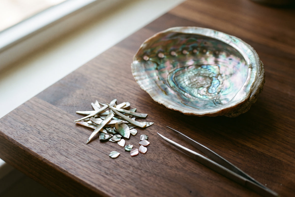 A mother-of-pearl shell beside thin cut slivers and tweezers on a walnut bench