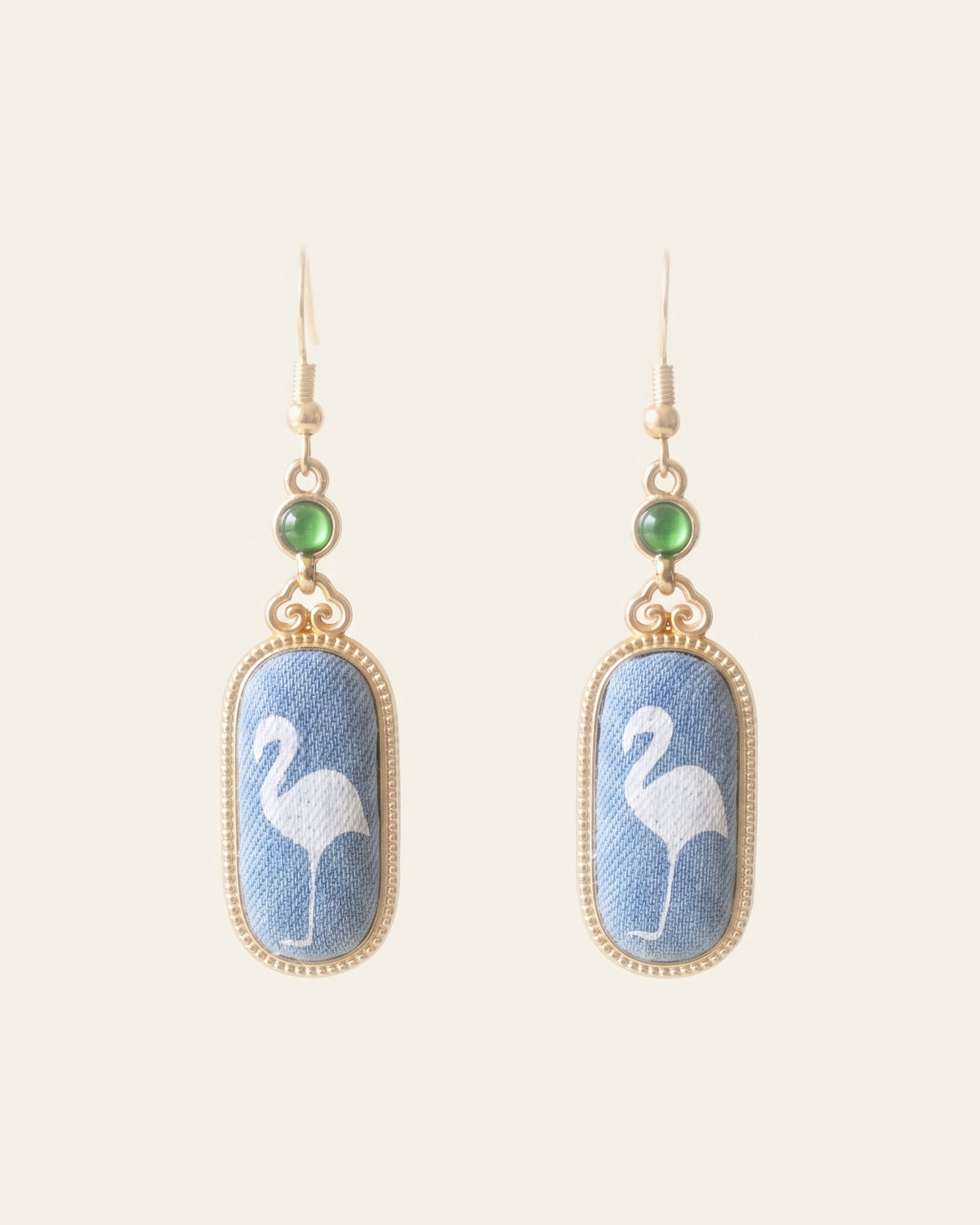 Pair of drop earrings with oval blue brocade panels showing a white flamingo, in beaded gold-tone frames below small green cabochons, on an ivory background