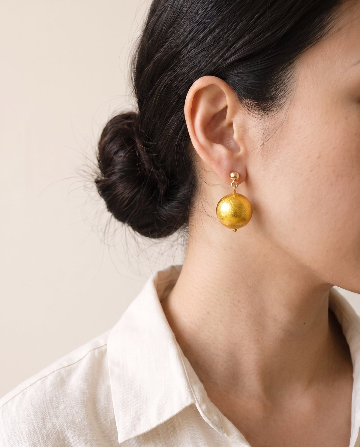 Gold leaf lacquer ball earring hanging from a small gold ball stud, worn with a linen shirt