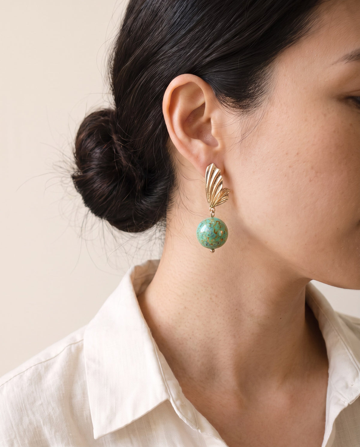 Turquoise Xipi lacquer bead earring hanging from a fluted gold wing stud, worn with a linen shirt
