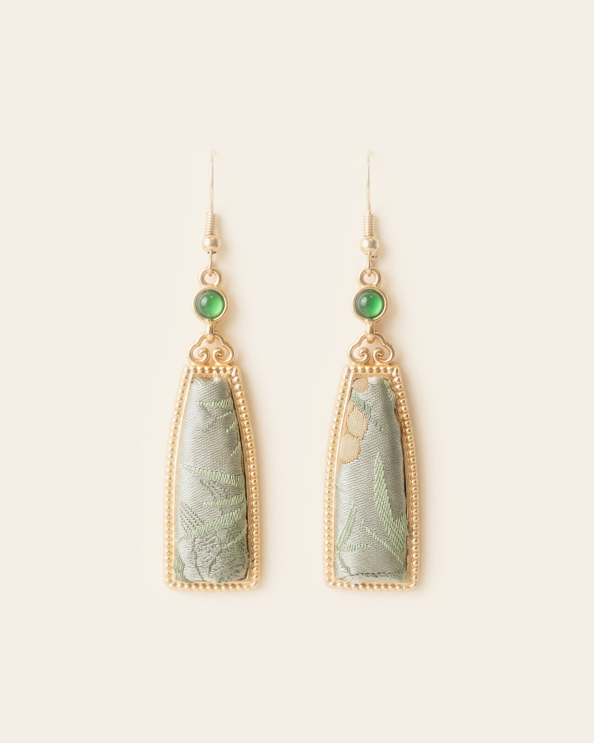 Pair of drop earrings: tapered panels of sage green Song brocade with a leaf pattern in beaded gold-tone Miao silver frames, below scroll links and round green cabochons, on gold-tone hook ear wires, on an ivory background.