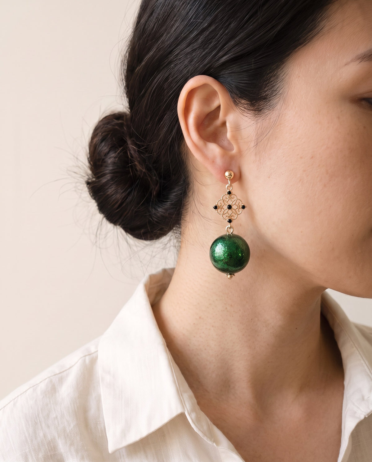 Emerald shimmer lacquer sphere earring below a gold filigree diamond set with five black stones, on a gold ball stud, worn with a linen shirt