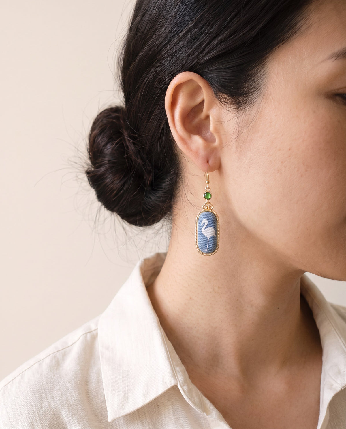 Blue Song brocade drop earring with a white flamingo in a gold-tone oval frame and green stone, worn with a linen shirt