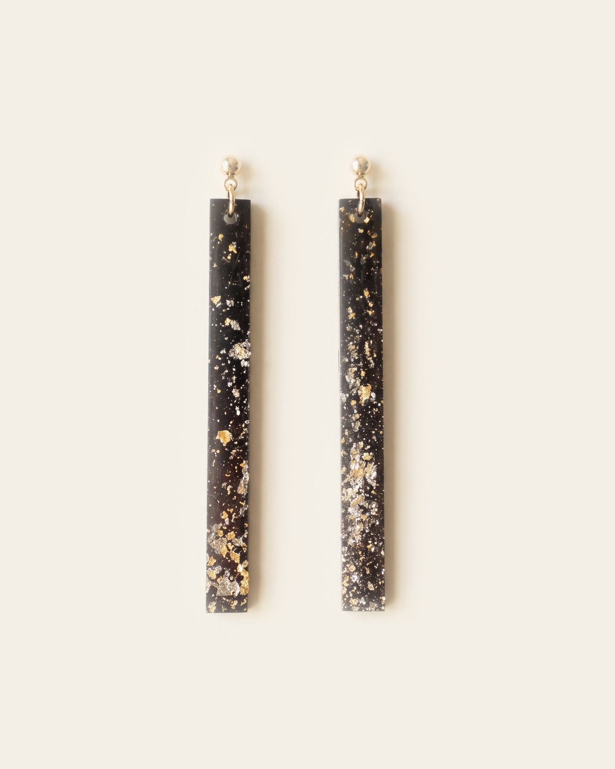 Pair of narrow black lacquer bar drop earrings flecked with gold flakes, hanging from gold ball studs, on an ivory background