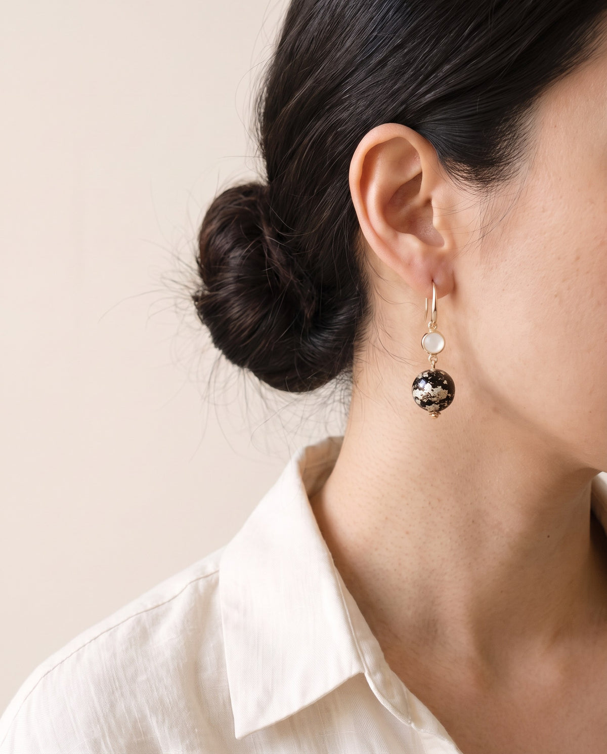 Black lacquer sphere with pale silvery leaf below a white mother-of-pearl cabochon in a gold bezel, on a gold French hook, worn with a linen shirt