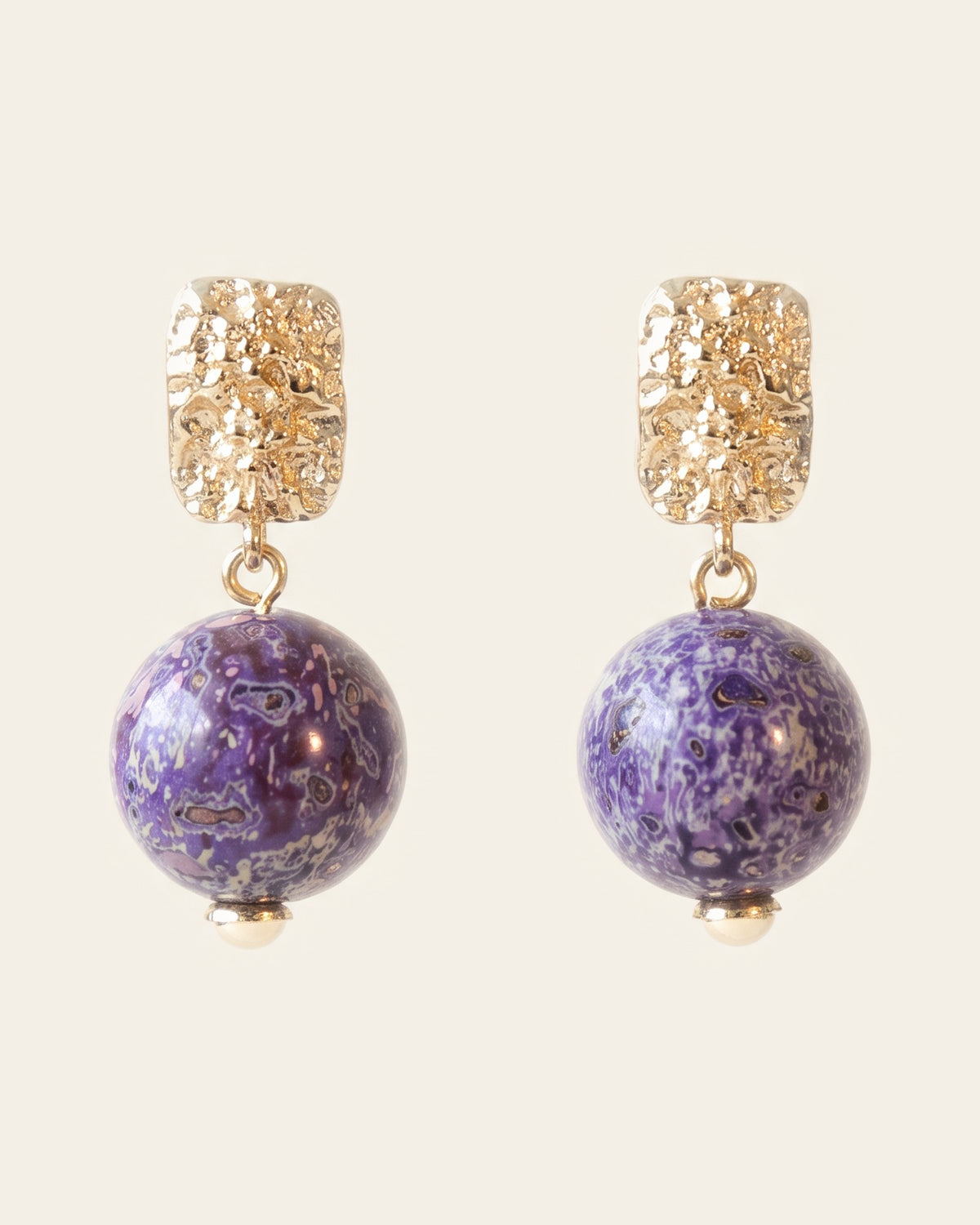 Pair of purple xipi lacquer sphere earrings marbled with lilac and grey, hanging from textured rectangular gold studs, on an ivory background