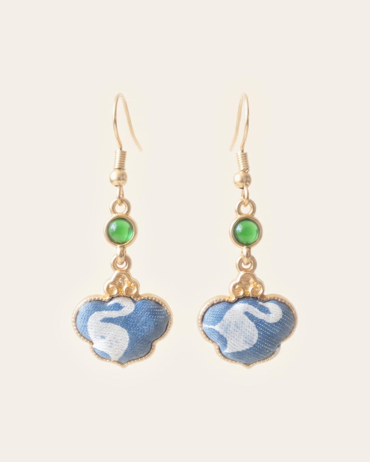 Pair of drop earrings with cloud-shaped blue brocade panels showing a white swan, in gold-tone frames below small green cabochons, on an ivory background