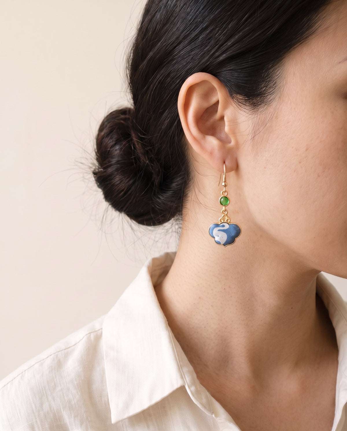 Blue Song brocade cloud earring with a white swan motif and a green cabochon, gold-tone hook, worn with a linen shirt