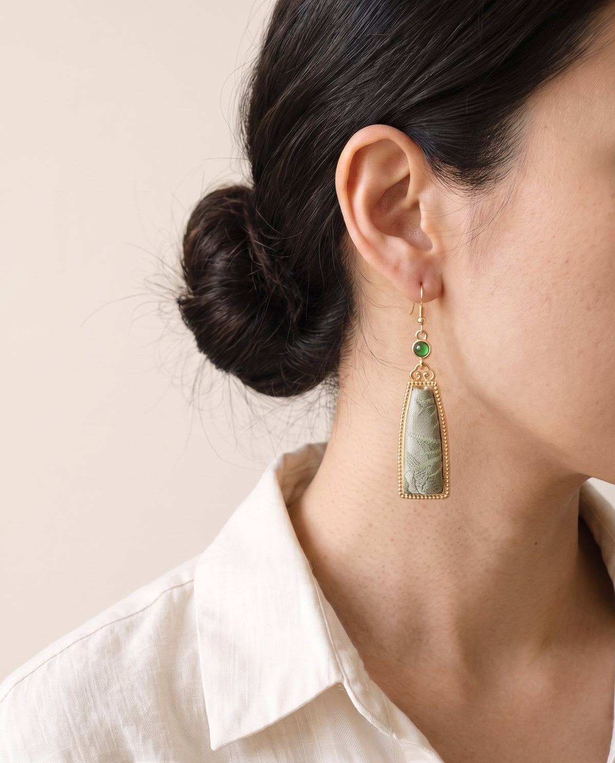 Sage green Song brocade leaf drop earring in a beaded gold-tone frame with a green stone, worn with a linen shirt