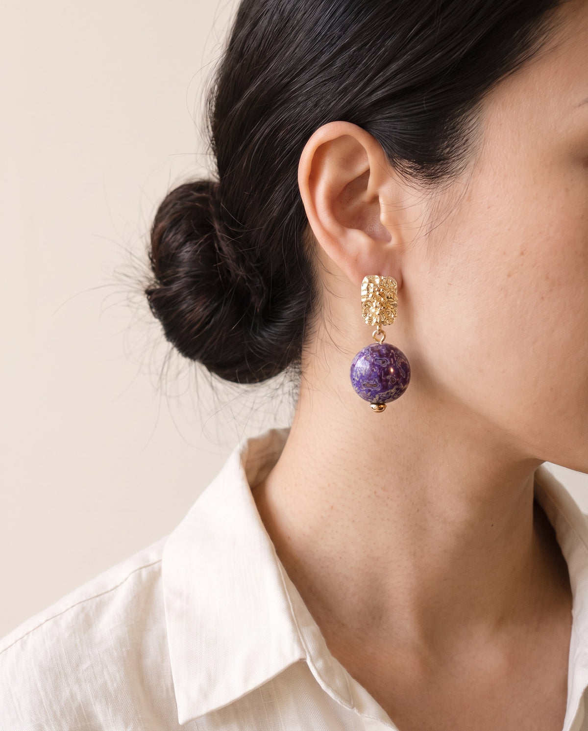 Purple Xipi lacquer bead earring hanging from a hammered gold rectangular stud, worn with a linen shirt