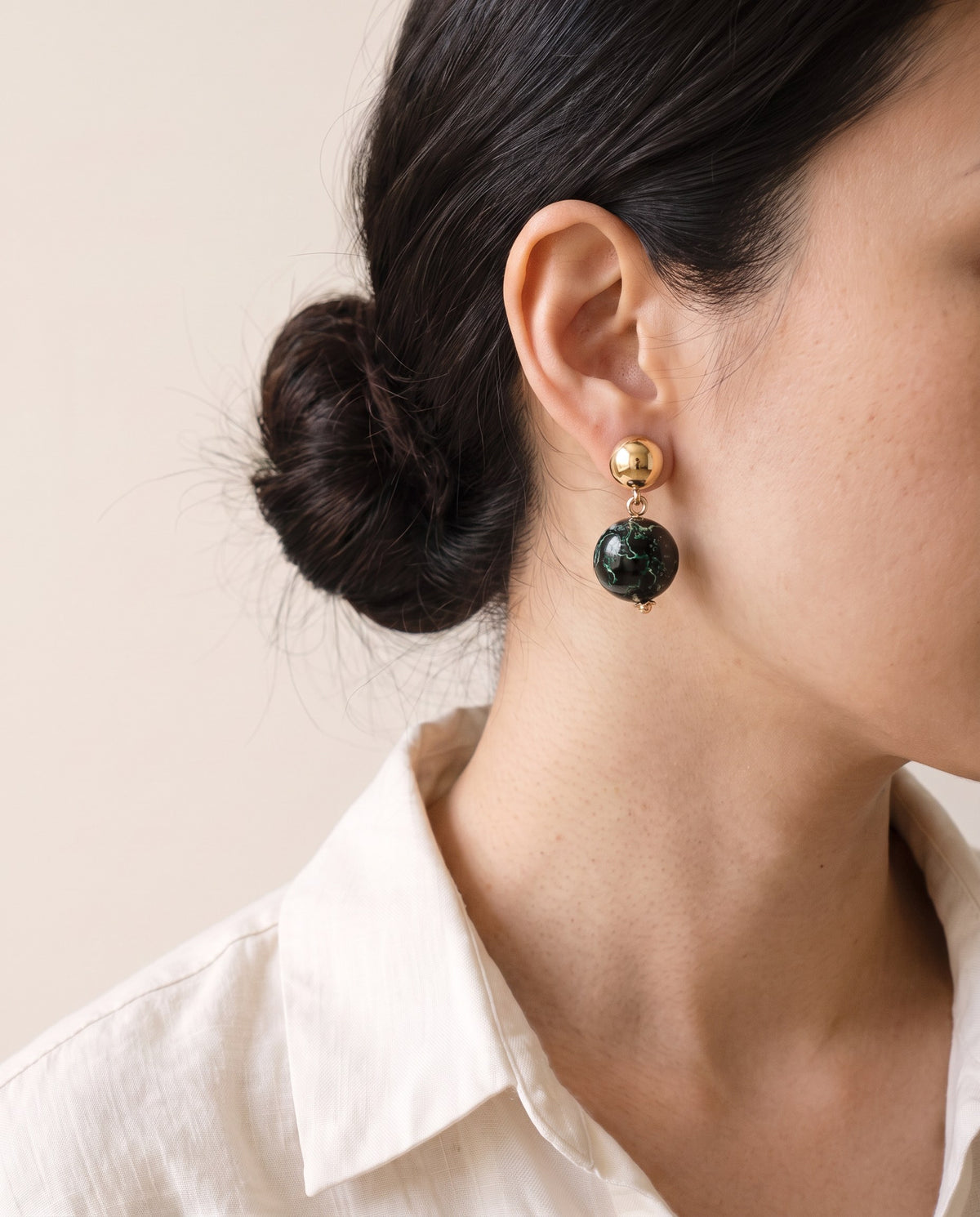 Black and green xipi lacquer bead earring hanging from a polished gold dome stud, worn with a linen shirt
