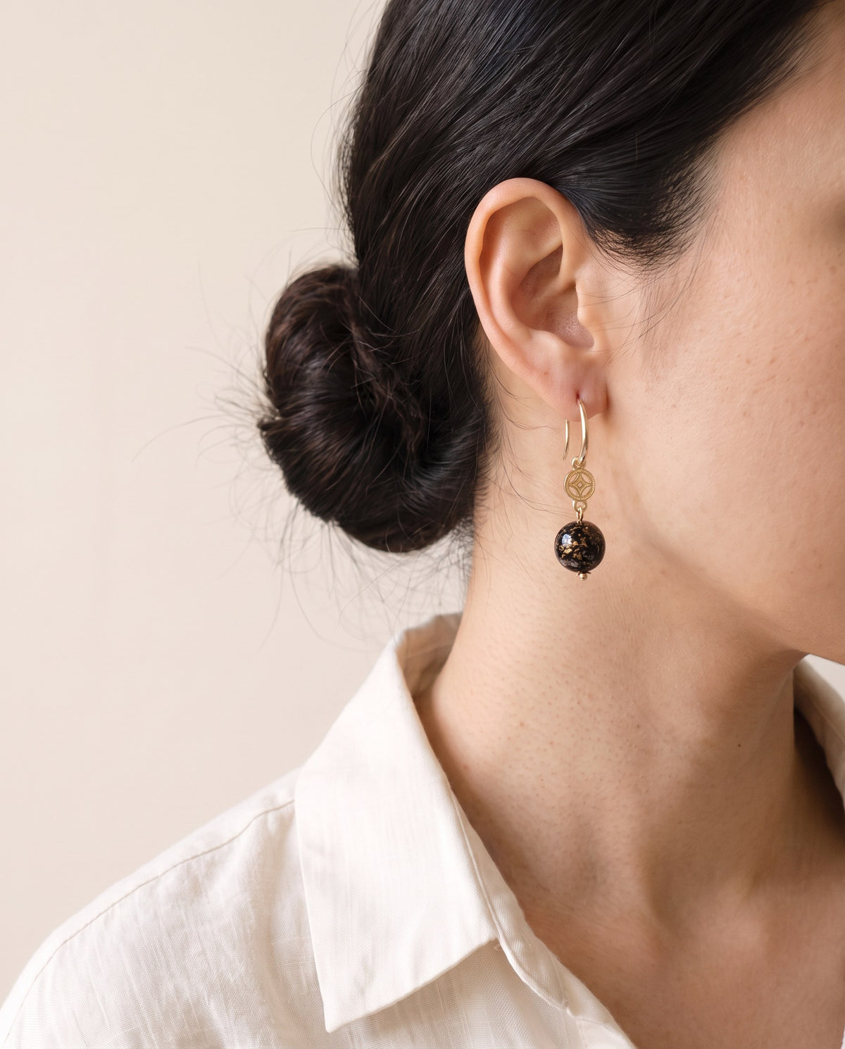 Black xipi lacquer sphere with gold flecks below a gold coin-flower charm on a gold hook, worn with a linen shirt
