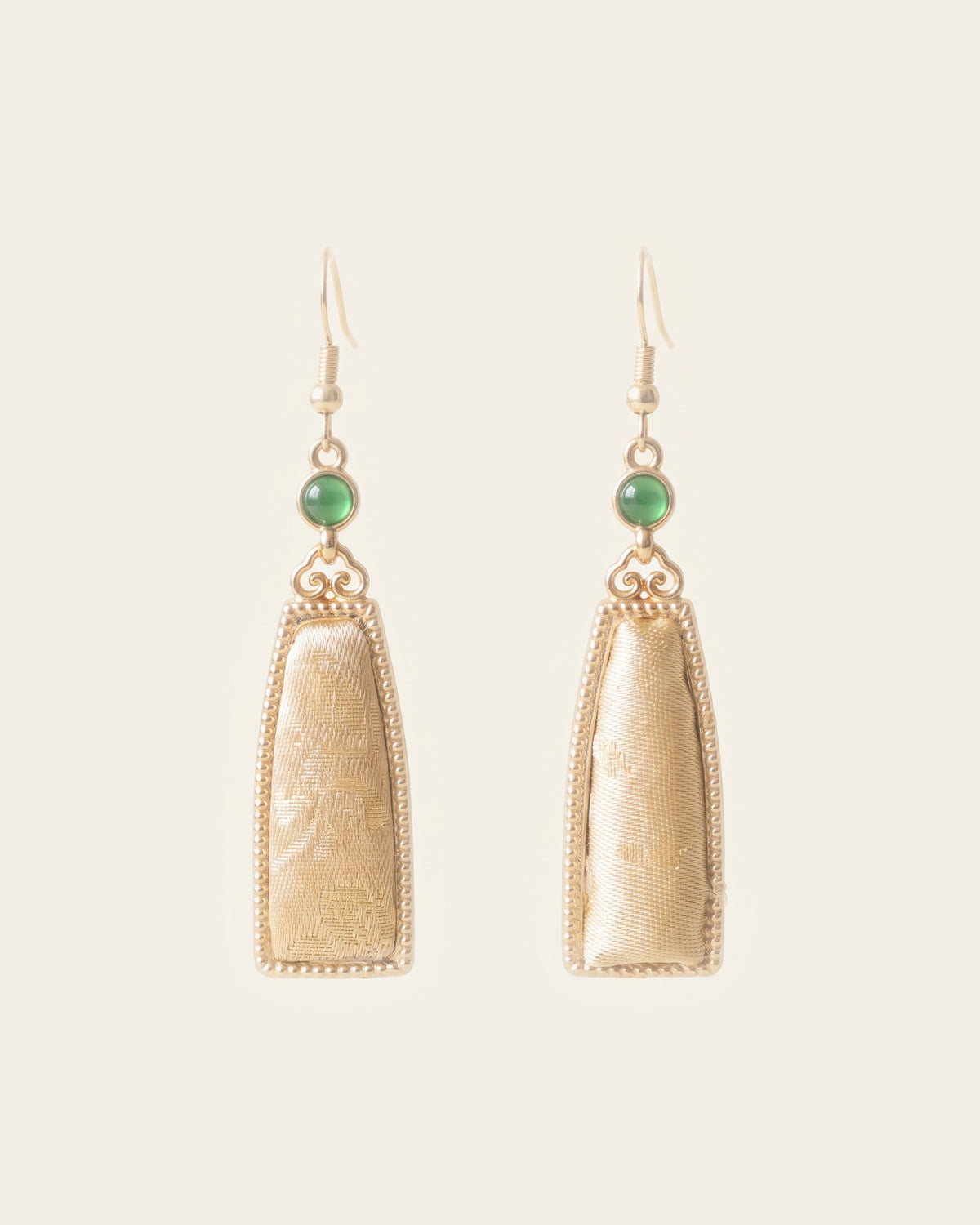 Pair of drop earrings: tapered panels of pale champagne Song brocade in beaded gold-tone Miao silver frames, below scroll links and round green cabochons, on gold-tone hook ear wires, on an ivory background.