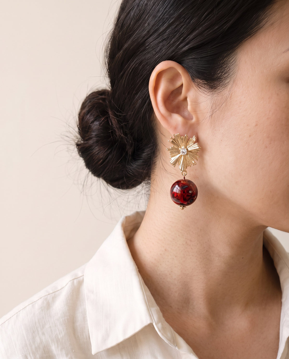 Red Xipi lacquer bead earring hanging from a gold starburst stud with a clear stone, worn with a linen shirt