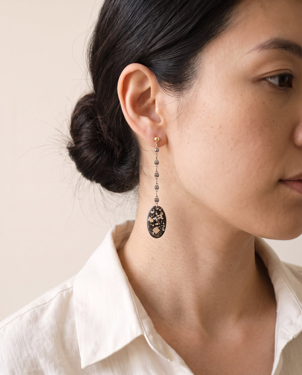 Black lacquer oval with gold and silver leaf and a small gold square, on a long gold-and-gunmetal bead chain from a ball stud, worn with a linen shirt