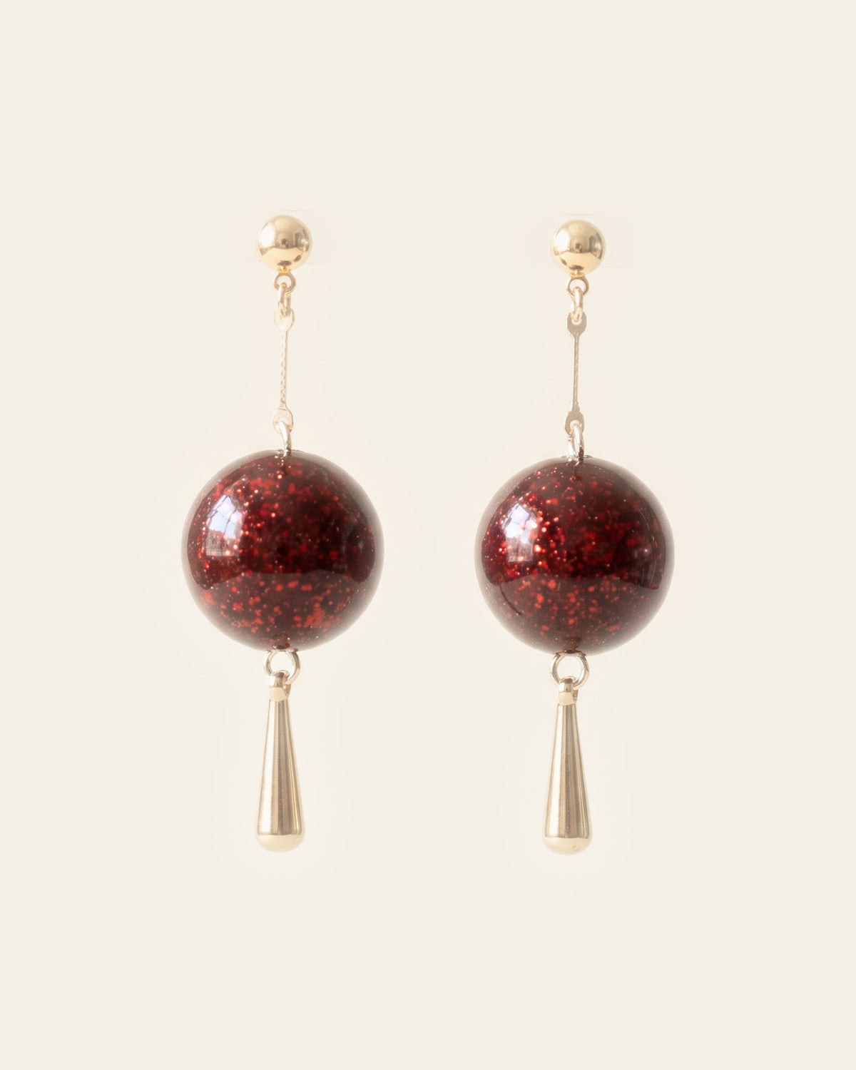 Pair of drop earrings with a glossy deep red flecked lacquer sphere and a gold teardrop below, on gold ball studs against an ivory background