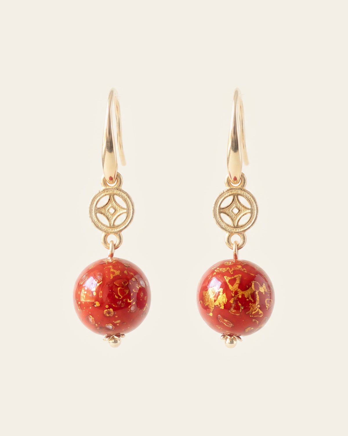 Pair of drop earrings with gold ear hooks, openwork gold coin charms and red lacquer spheres flecked with gold, on an ivory background