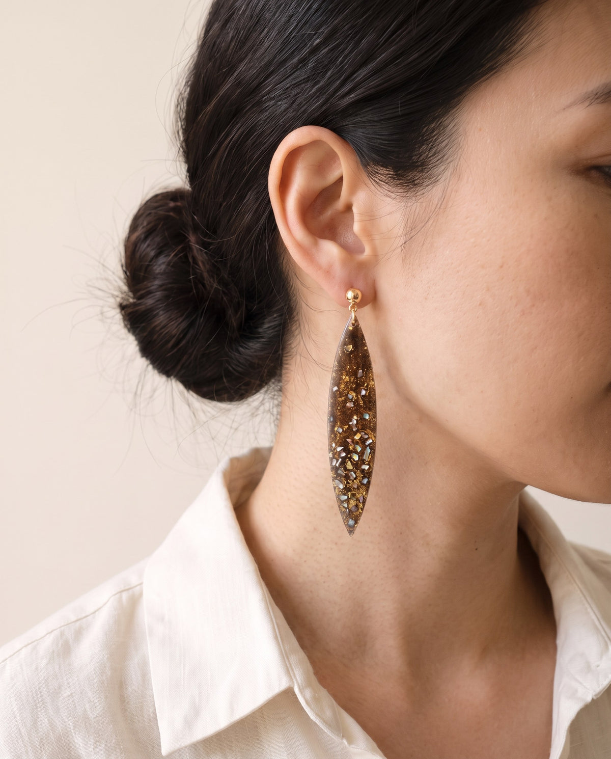 Long brown lacquer leaf earring flecked with gold leaf and mother-of-pearl on a gold ball stud, worn with a linen shirt