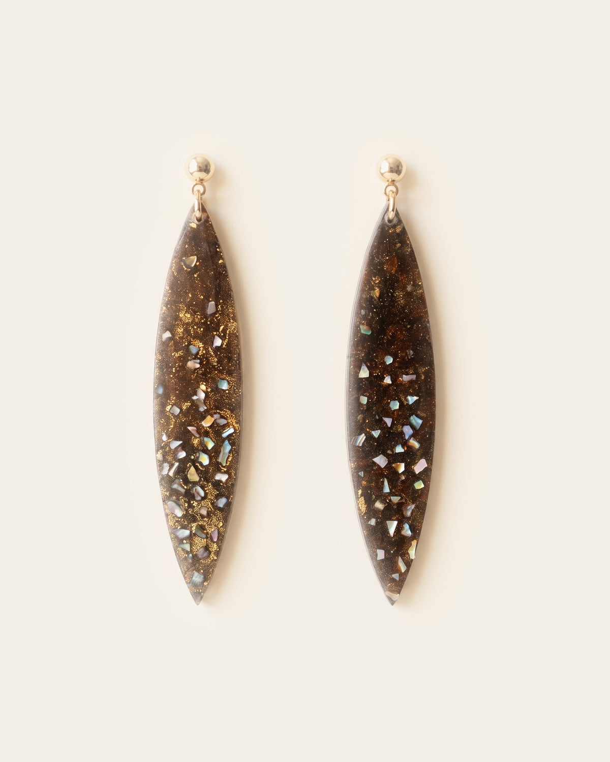 Pair of long leaf-shaped drop earrings in dark brown lacquer with gold leaf and mother-of-pearl flecks, hanging from gold ball studs on an ivory background