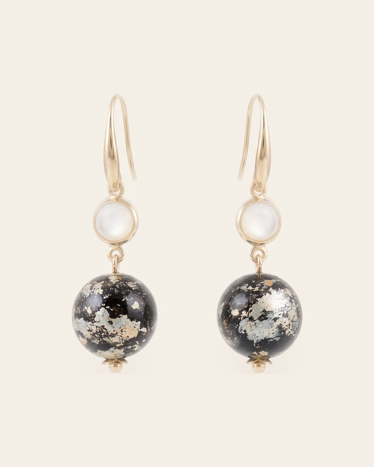 Pair of drop earrings: near-black lacquer spheres with pale silvery-gold leaf below white cabochons in gold bezels, on gold ear hooks, ivory background