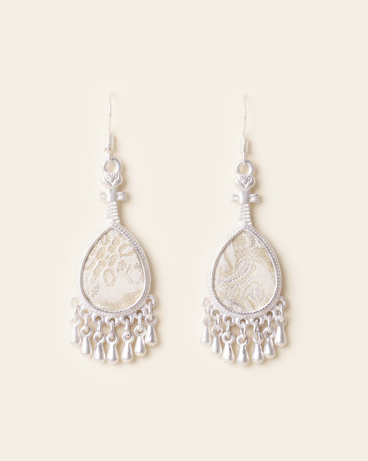 Pair of silver-tone pipa-shaped drop earrings set with ivory and pale gold Song brocade and finished with a fringe of small silver teardrops, on silver-tone hooks against an ivory background