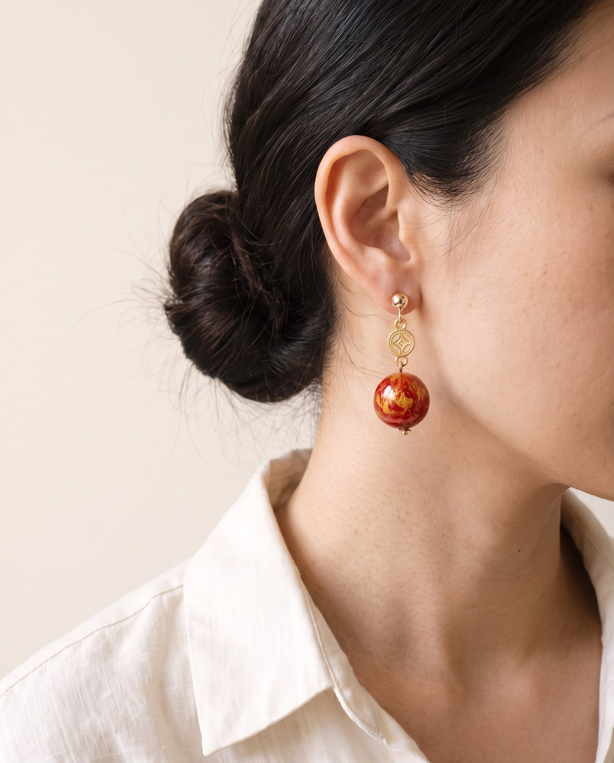 Gold-marbled red lacquer bead earring with an openwork gold coin charm on a small ball stud, worn with a linen shirt