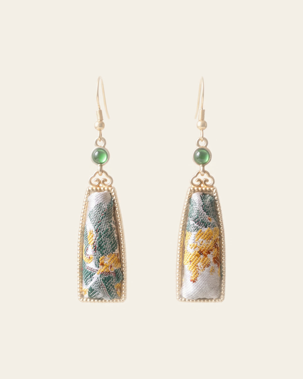 Pair of long tapered drop earrings with silver-white Song brocade panels patterned in green leaves and a yellow flower, in gold-tone frames with small green cabochons, on gold-tone hooks against an ivory background