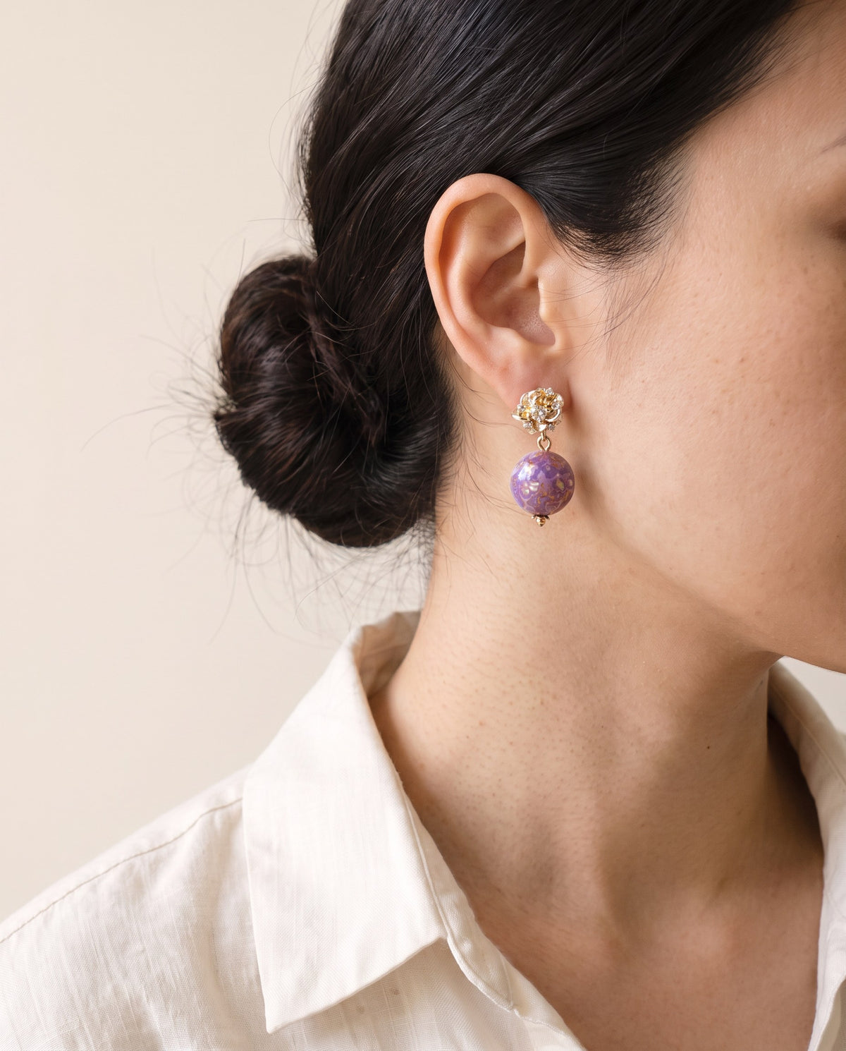 Lilac marbled lacquer bead earring with mother-of-pearl flecks on a gold flower stud set with clear stones, worn with a linen shirt