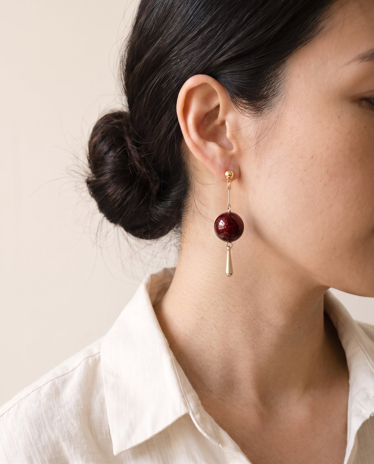 Red flecked lacquer sphere earring on a gold ball stud with a slim bar chain and gold teardrop drop, worn with a linen shirt