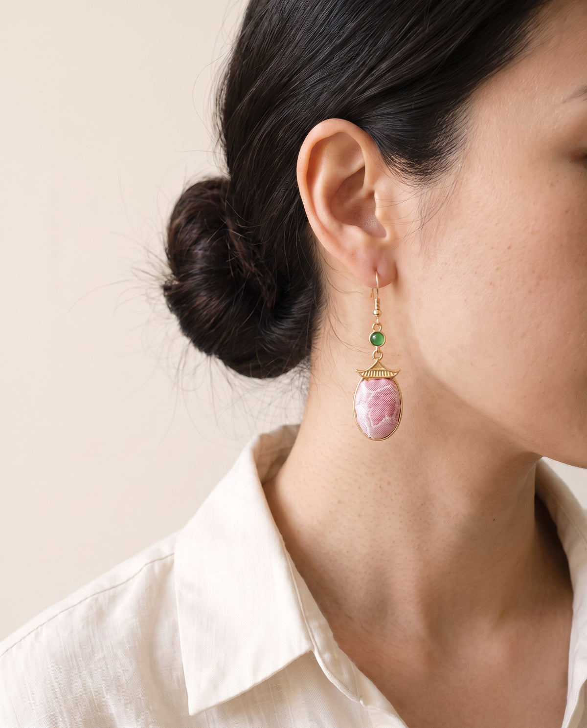 Pink Song brocade oval earring under a gold pagoda-roof cap with a green cabochon, worn with a linen shirt