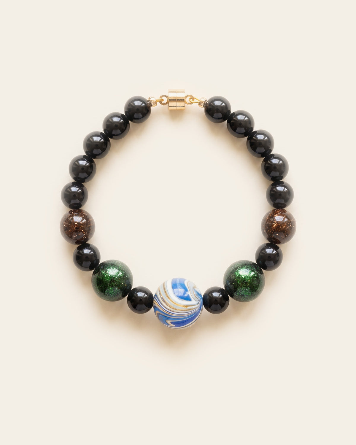 Black lacquer bead bracelet with a blue and white marbled centre bead, green sparkle beads and translucent amber beads with copper flecks, and a gold magnetic clasp, laid flat on an ivory background