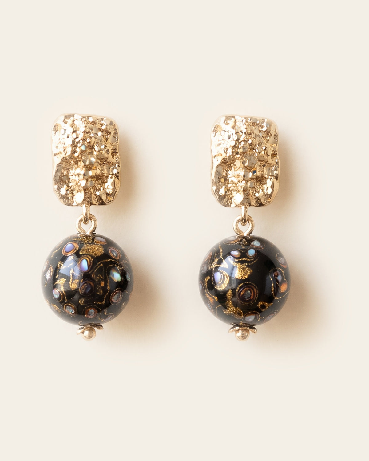 Pair of black xipi lacquer sphere earrings with gold swirls and mother-of-pearl inlay, hanging from textured rectangular gold studs, on an ivory background