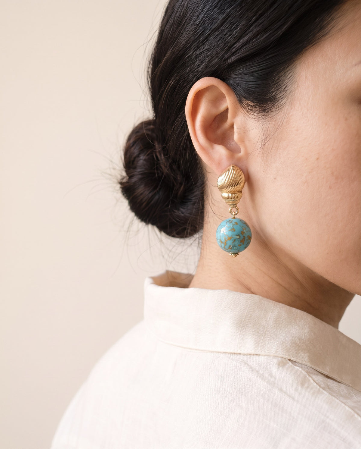 Turquoise Xipi lacquer bead earring below a ribbed gold seashell stud, worn with a linen shirt