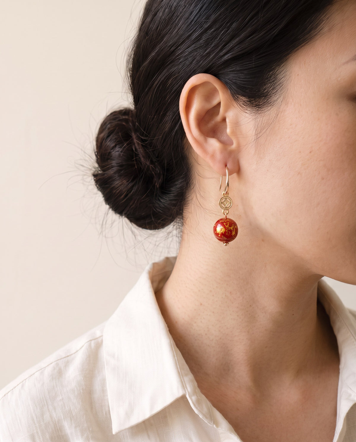Red Xipi lacquer bead with gold leaf below a gold coin charm on a hook, worn with a linen shirt