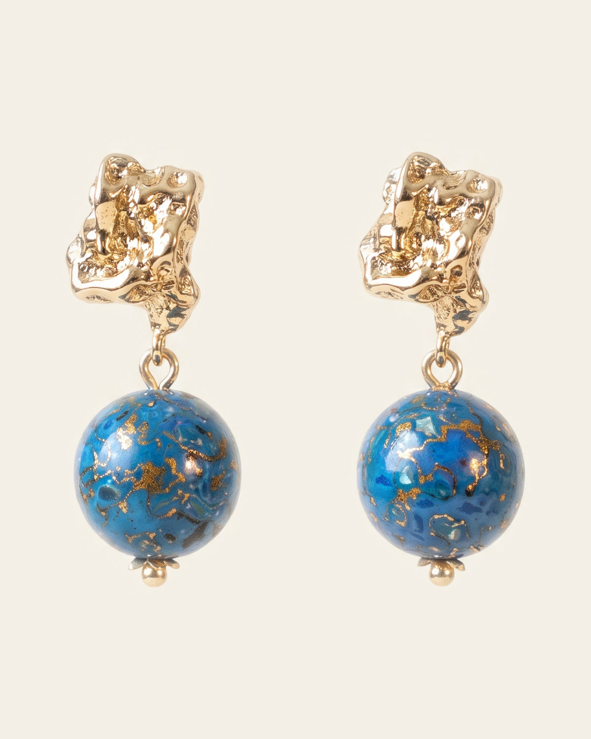 Pair of drop earrings: textured gold nugget post studs holding round deep blue xipi lacquer beads flecked with gold, on an ivory background.
