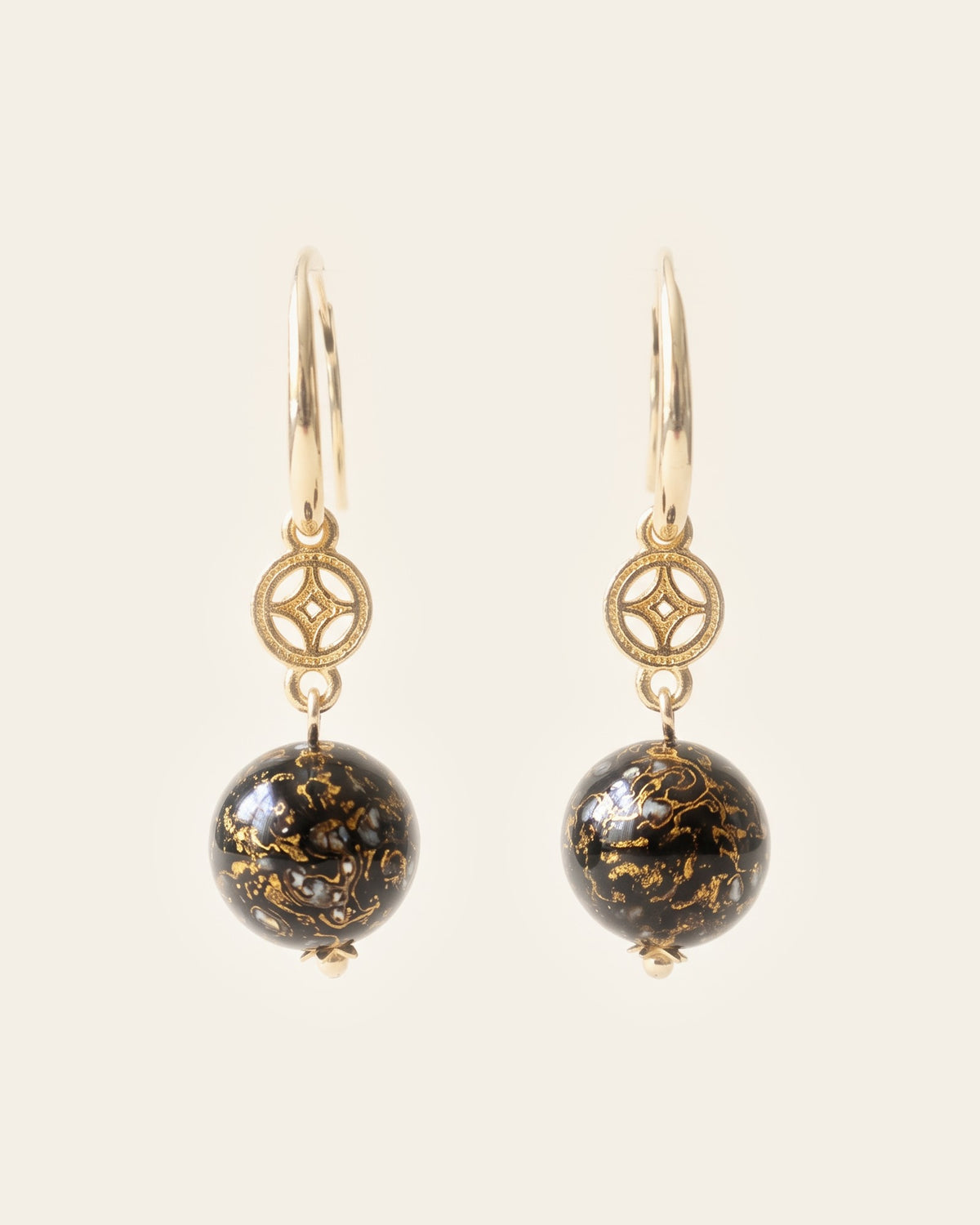 Pair of black xipi lacquer sphere earrings with gold marbling and mother-of-pearl flecks, hanging from 18k gold coin-flower charms on 14k gold ear hooks, on an ivory background