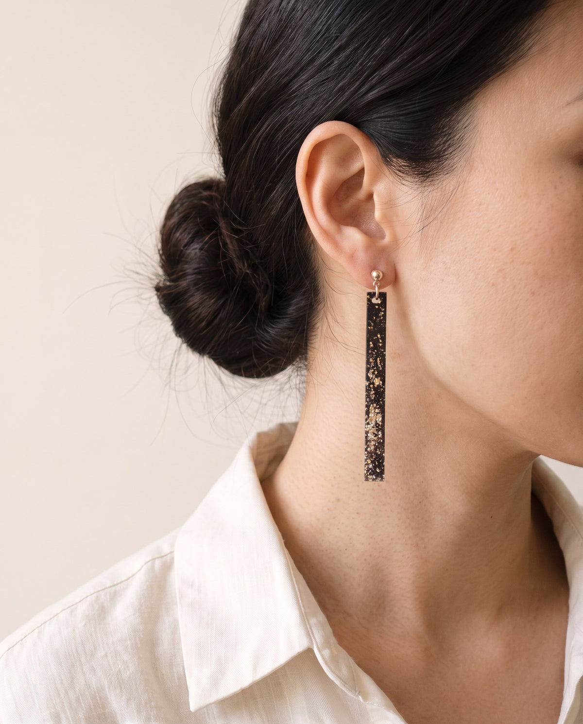 Long black lacquer bar earring scattered with gold and silver leaf, hanging from a gold ball stud, worn with a linen shirt