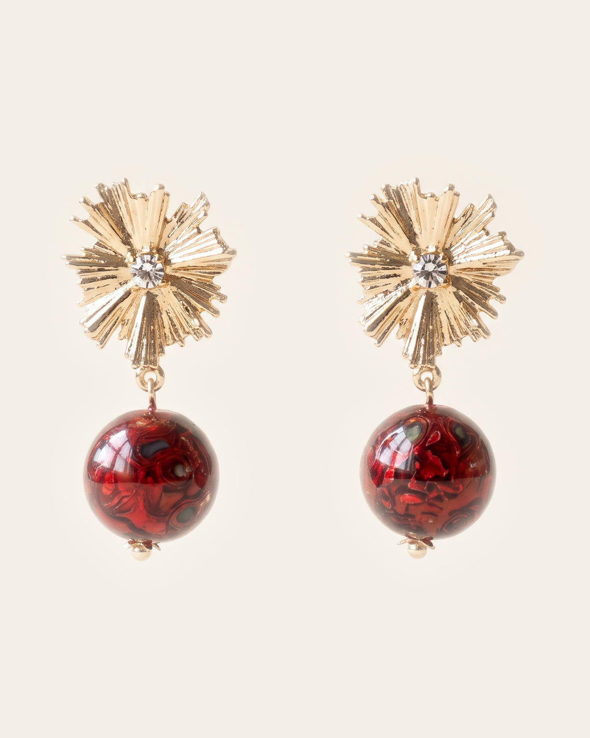 Pair of drop earrings: ribbed gold starburst post studs each set with a clear stone, holding round deep red xipi lacquer beads, on an ivory background.