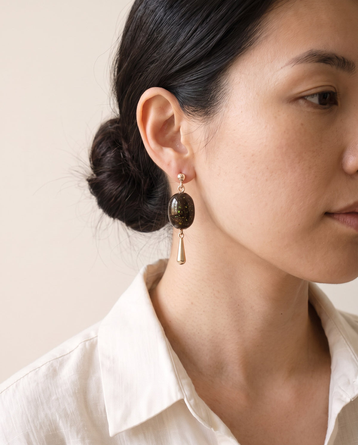 Brown lacquer oval earring flecked with gold and green, with a gold ball stud and gold teardrop drop, worn with a linen shirt